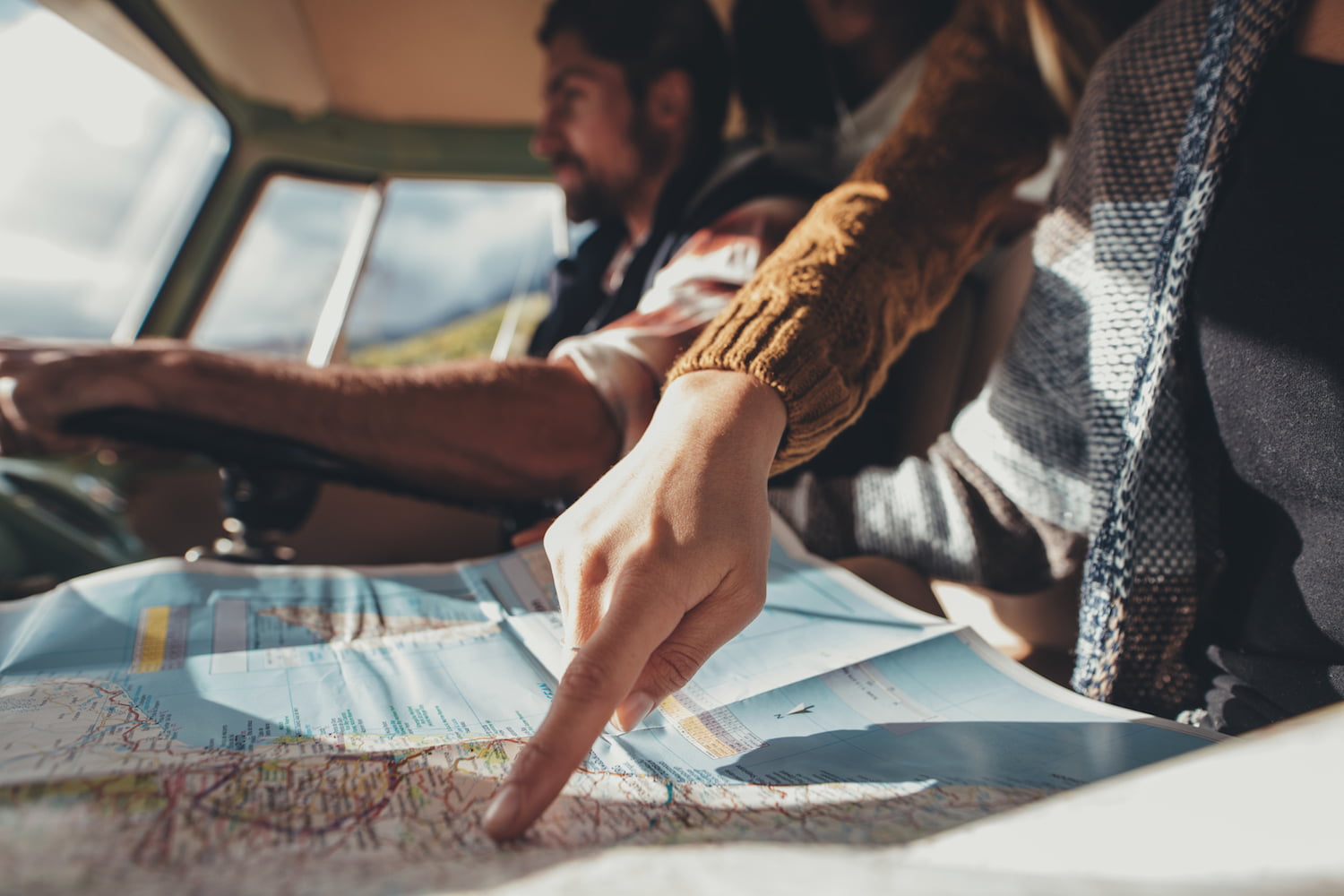 Friends on road trip looking at map for directions. Close up of female pointing at the map while man driving van in background.