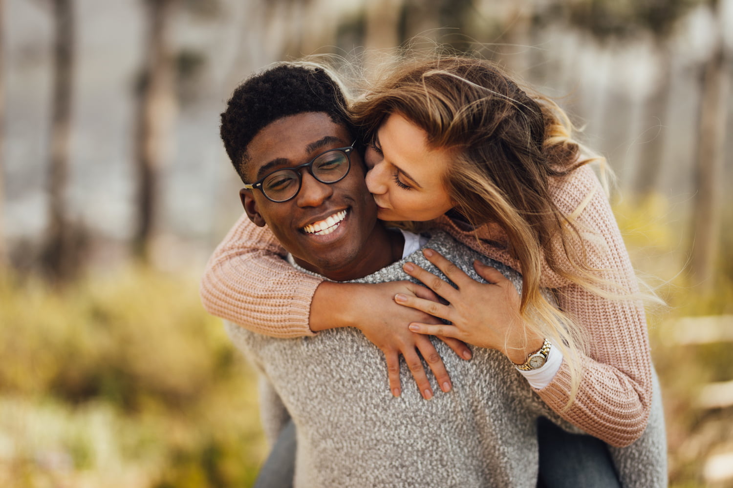 Handsome young man giving piggyback ride to his girlfriend. Couple having fun outdoors. Man carrying his girlfriend on his back, with woman kissing man on the cheek. (Handsome young man giving piggyback ride to his girlfriend. Couple having fun outdoo